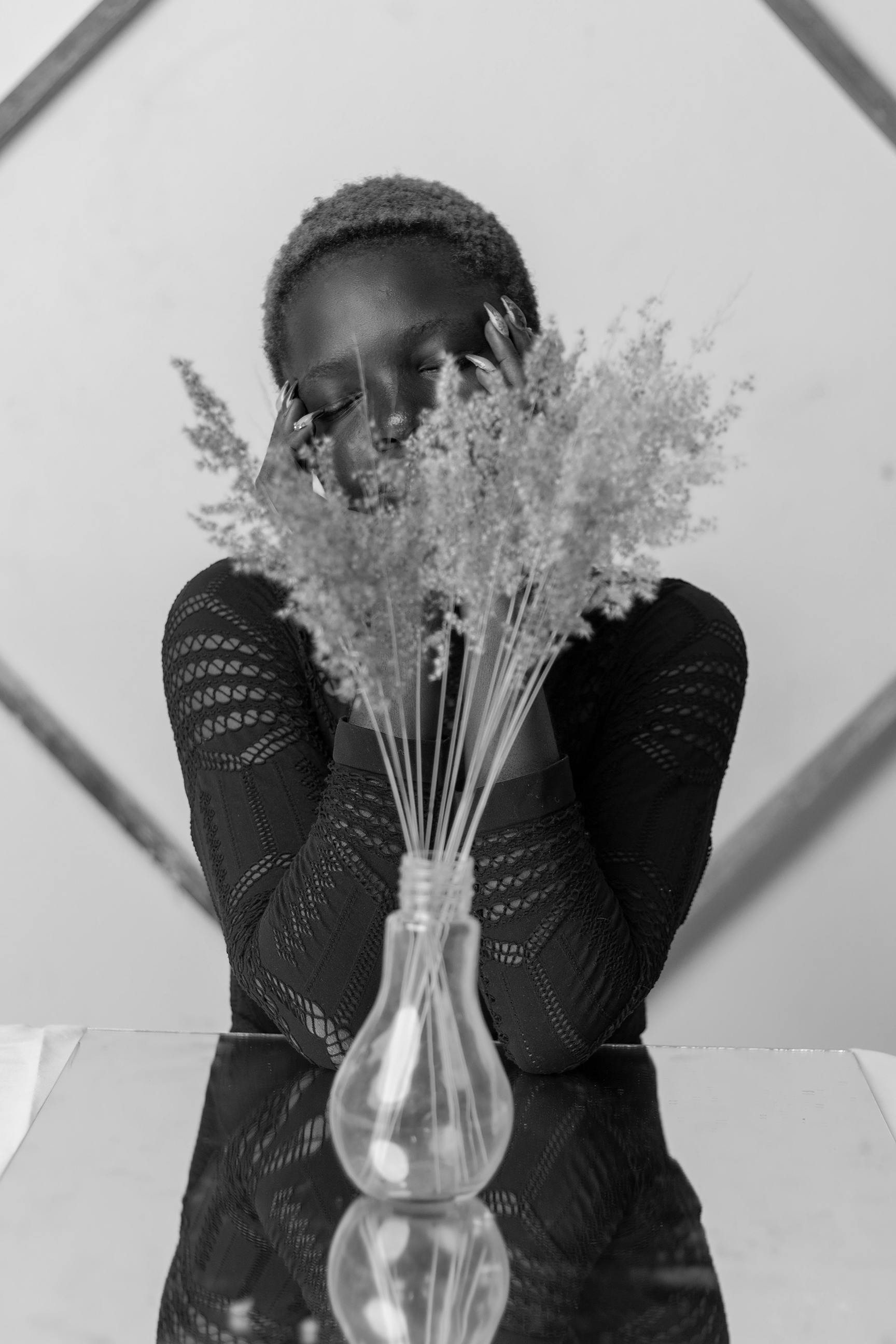 Black and white of melancholic young ethnic woman with closed eyes sitting at table with flowers vase placed on mirror
