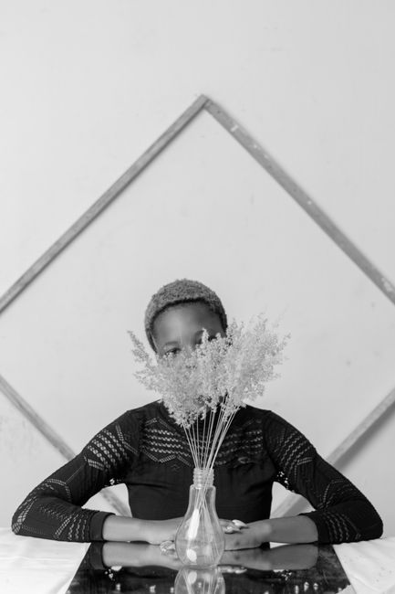 Black and white of unrecognizable young African American female in elegant dress resting at table with bunch of delicate flowers in vase against wall with geometric decor