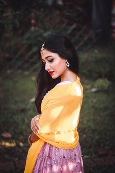 Side view of dreamy Asian female in vivid scarf and festive dress looking down on blurred background of green forest
