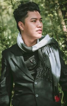 一位穿着西装、系着围巾的时尚亚洲男子，在郁郁葱葱的户外环境中凝望远方