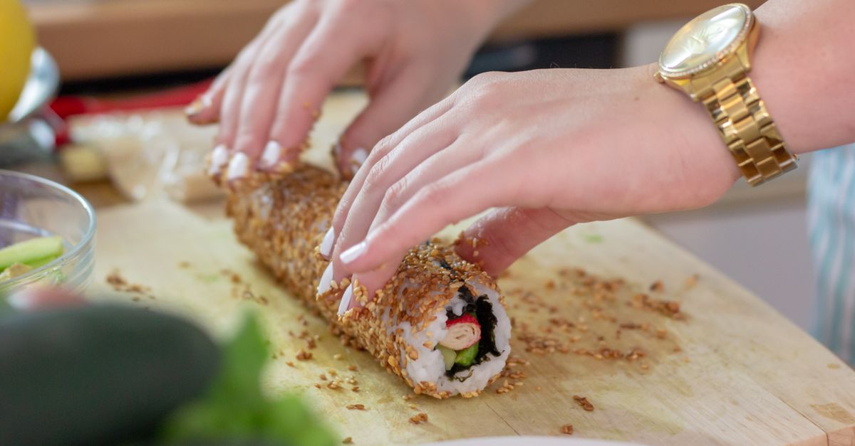 Hände rollen mit Sesam ummanteltes Sushi auf einem Holzbrett.