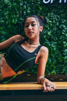 Chic young woman in a black tank top posing against a green leafy background with confidence.