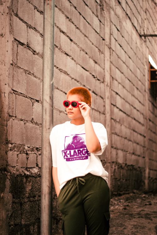 Free A fashionable woman with short hair poses in an alley, wearing red sunglasses and a stylish white shirt. Stock Photo