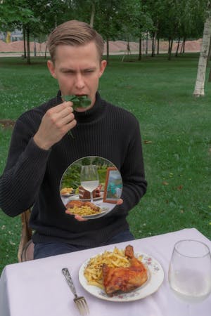 Male sitting at table on grassy ground with mirror in hand reflecting heavy meal while eating helathy salad in outdoor cafe