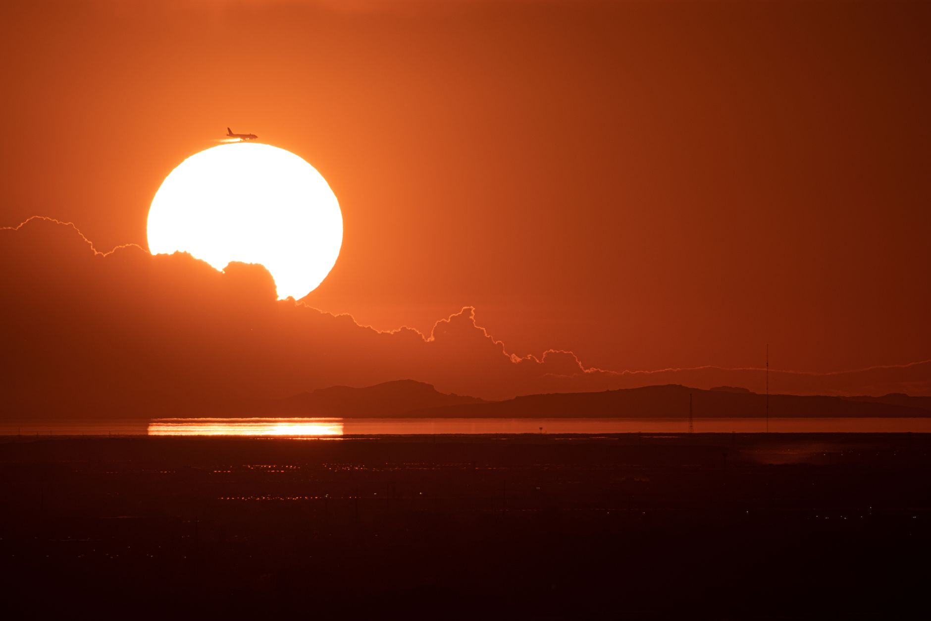 Silhouette of airplane passing giant sun at sunset over the ocean, creating dramatic sky and reflections.