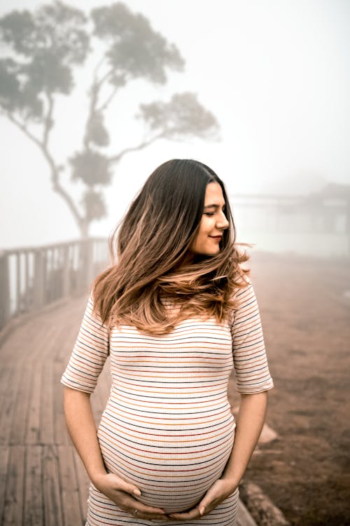 Calm young pregnant female touching belly and standing on wooden walkway on nature in foggy day