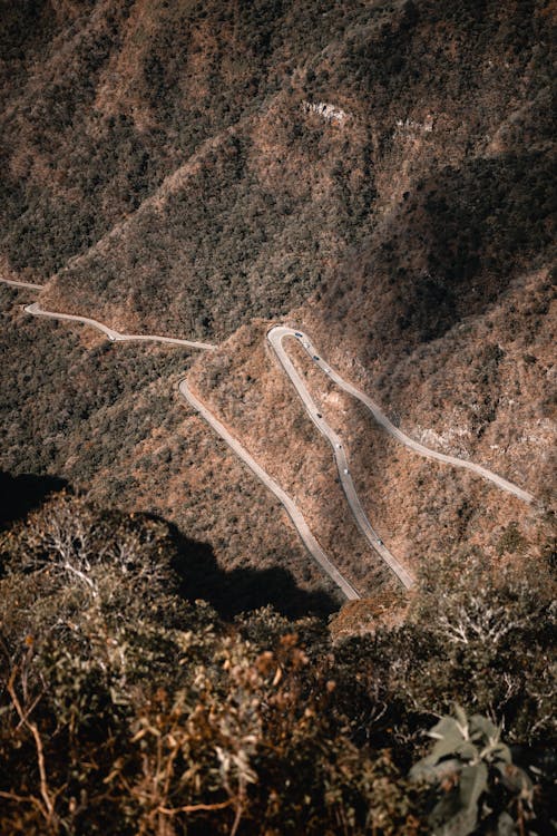 From above of cars driving on asphalt roadway running through rocky slopes covered with plants in highland area