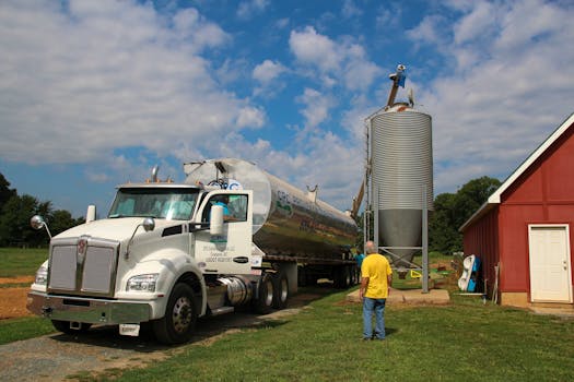 A fuel truck delivers to a silo on a farm under a blue sky.