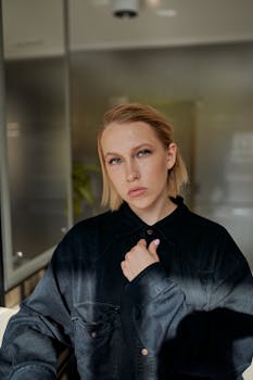 Serene young woman with short blond hair poses indoors in soft light, wearing dark clothing.