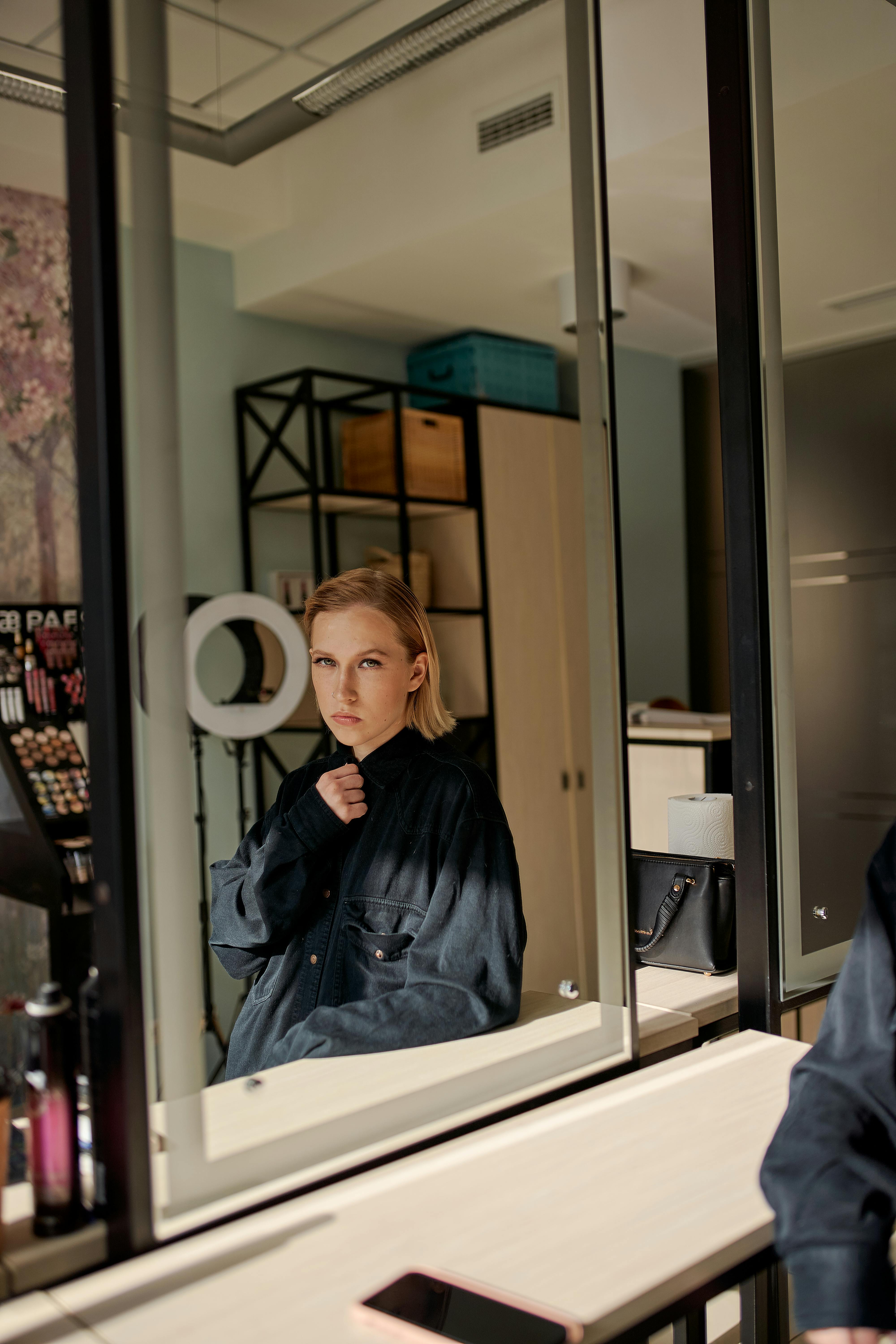 Crop young focused female model in black apparel looking in mirror while sitting at table in beauty salon