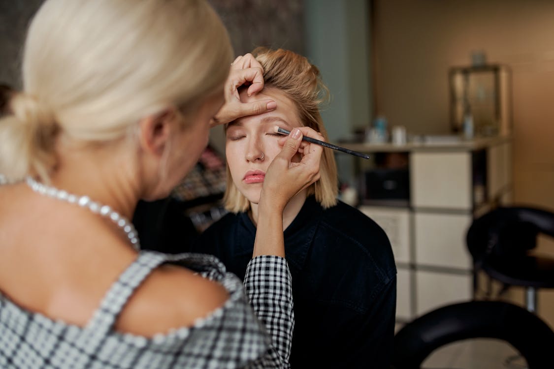 Free Crop anonymous beauty master doing makeup to female model with closed eyes using applicator in salon Stock Photo