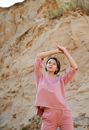 Young contemplative woman in trendy clothes standing with raised arms near dry rock while looking up