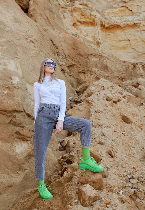 Young stylish female in bright apparel and contemporary sunglasses standing on dry rock in daylight