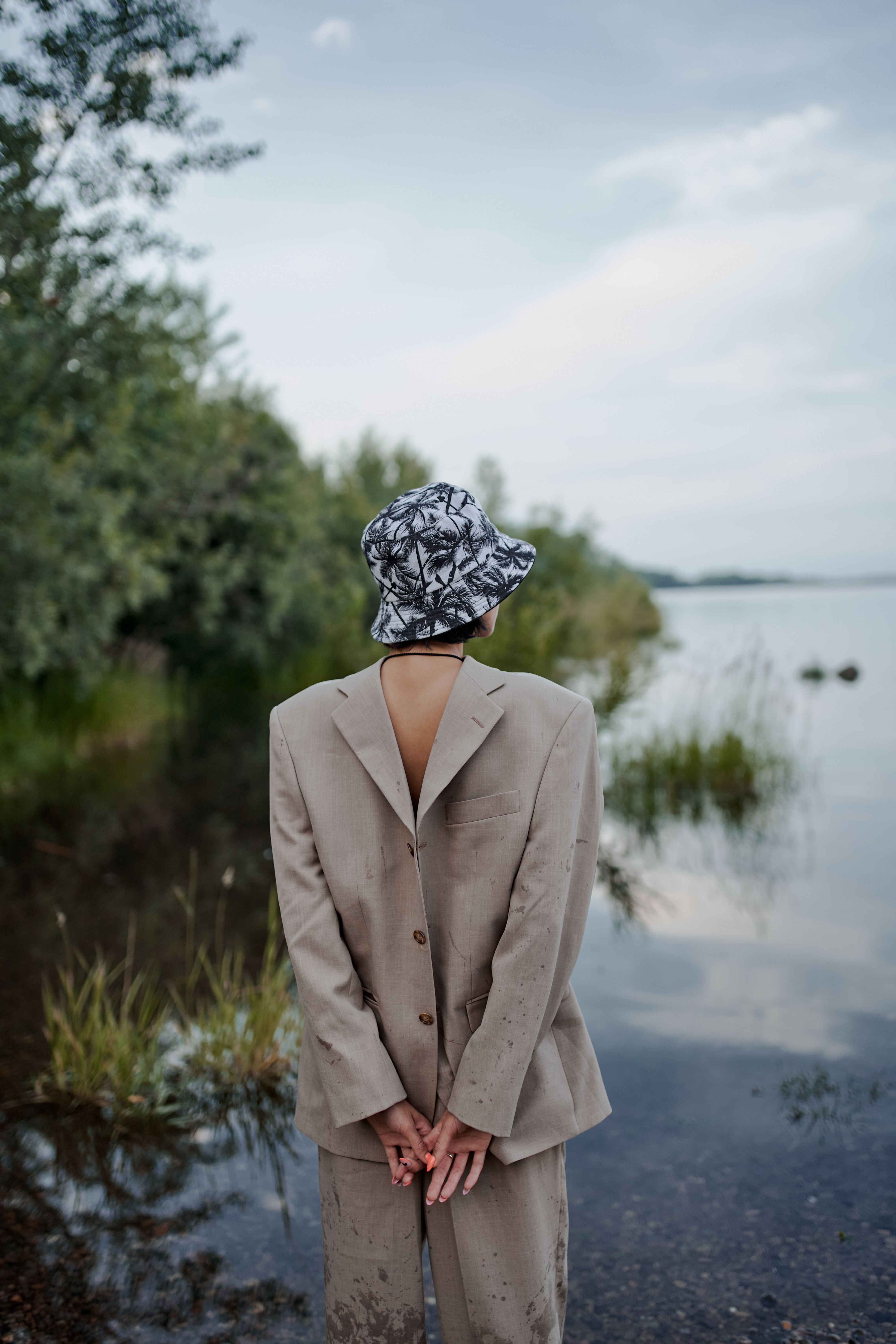 一位身穿夹克的匿名女性背对镜头，双手背在身后，凝视着乡间的河流
