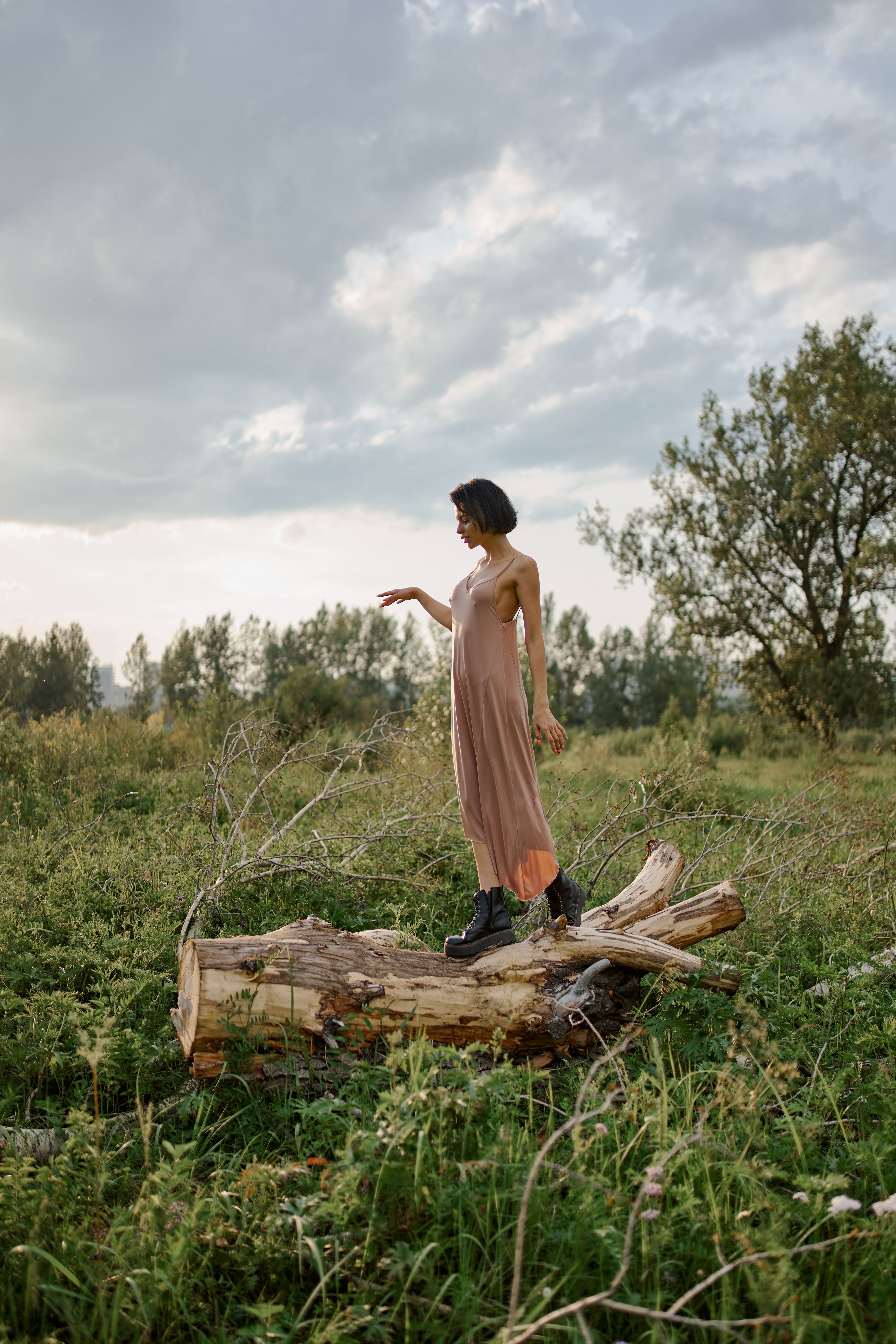 一名身穿太阳裙的苗条女子，在多云的天空下，优雅地站在郁郁葱葱草地上的一根木头上