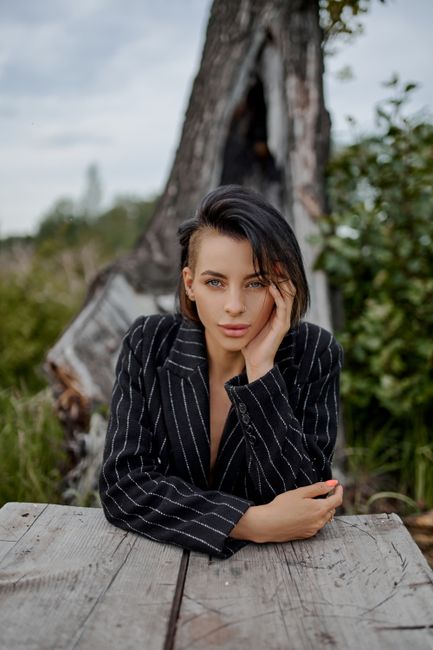 Young concentrated trendy lady with modern haircut looking at camera at wooden table under cloudy sky