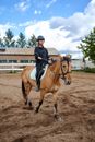Full body of serious young female jockey in helmet and gloves keeping bridle while riding obedient bay dun horse in paddock