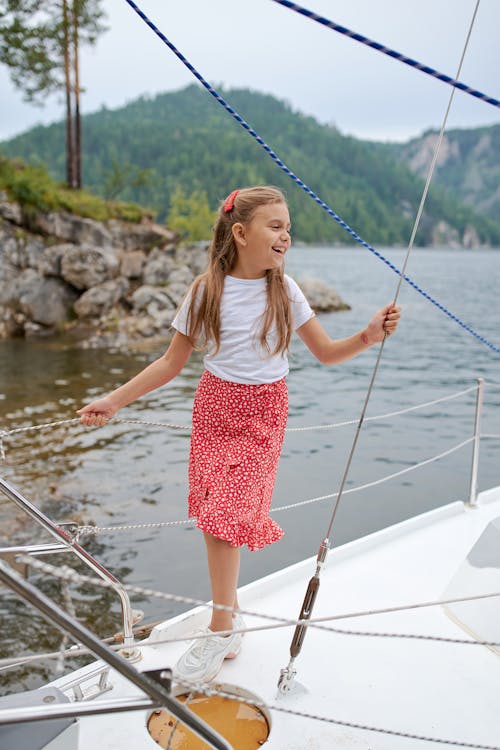 Full body of happy child in summer clothes standing on boat and laughing while spending time in nature