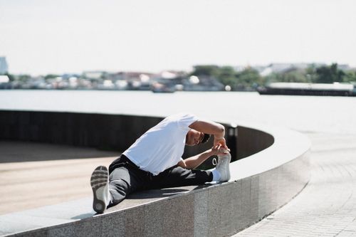 Free Young ethnic male in sportswear stretching legs while sitting on rounded fence near pavement in town Stock Photo