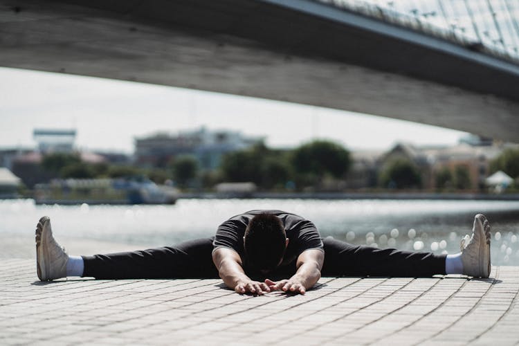 Faceless Man Stretching Legs On Embankment Near City River