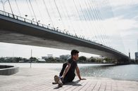 一名年轻的少数族裔男子在河流和桥梁附近练习瑜伽，目光却移向别处