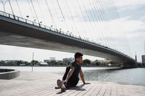 Free Young ethnic guy practicing yoga performing Seated Twist pose near river and bridge while looking away Stock Photo