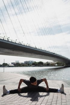 河岸边，一名面目全非的年轻少数族裔男子在多云的天空下伸腿，他的影子投在河边