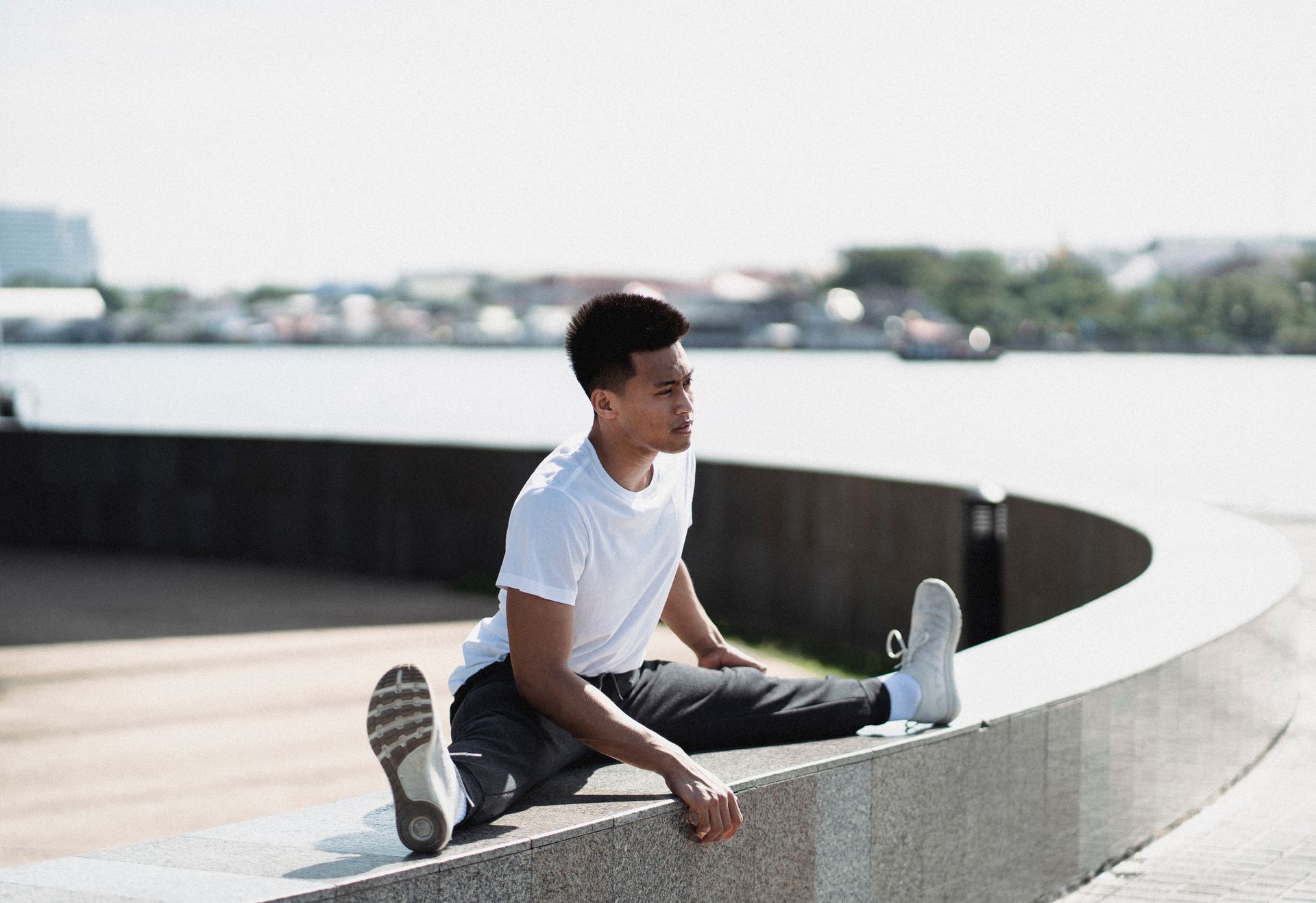 An Asian man performing a split stretch on an urban embankment beside a river, emphasizing flexibility and fitness.