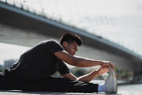 Flexible young Asian sportsman in black sportswear doing split exercise and stretching legs while warming up on river shore near urban bridge