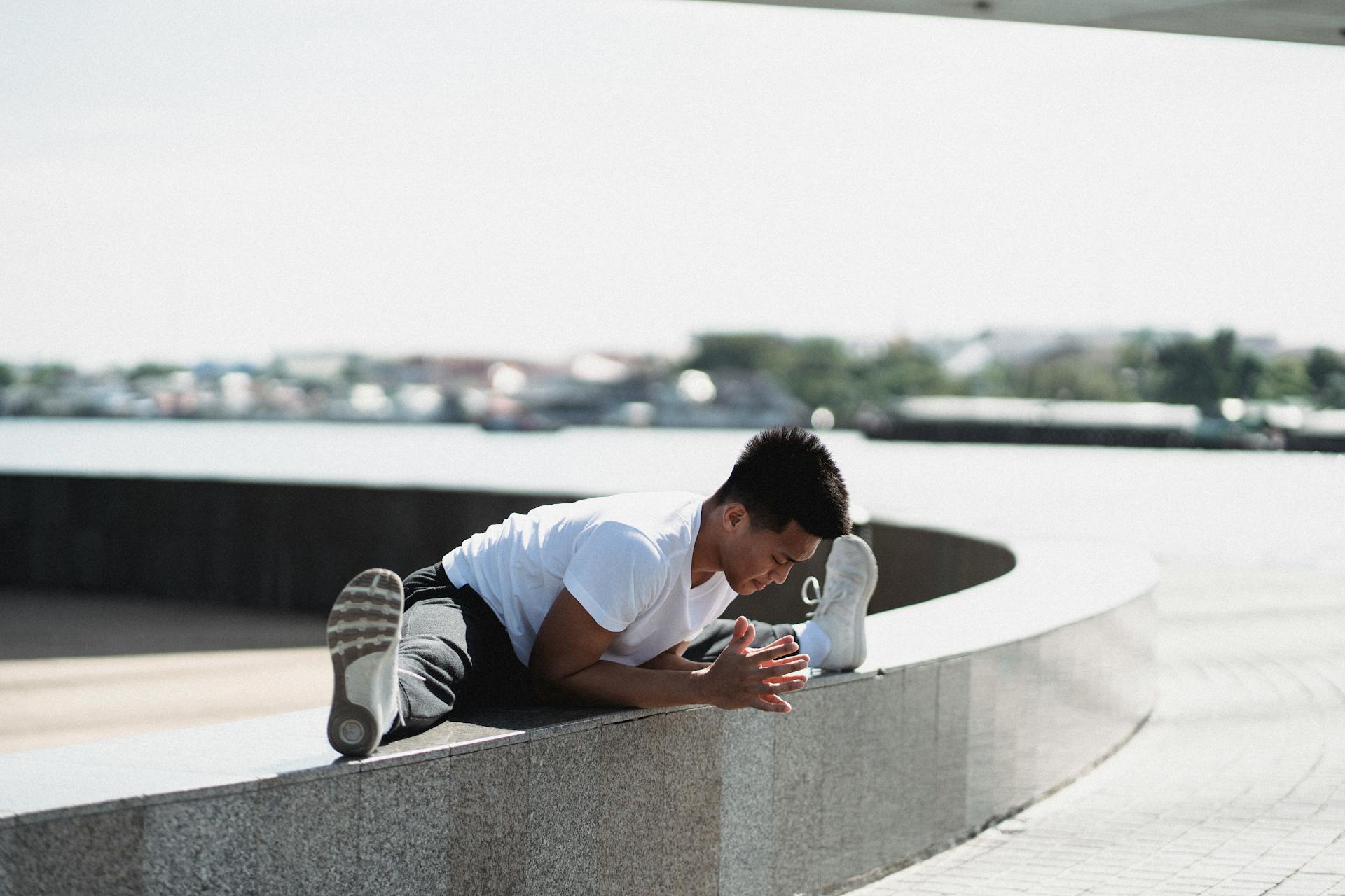 Full body sporty young Asian male in activewear doing split and bending body towards while warming up on embankment border on sunny weather