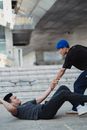 Side view young Asian man wearing street style outfit helping fallen friend to get up from ground