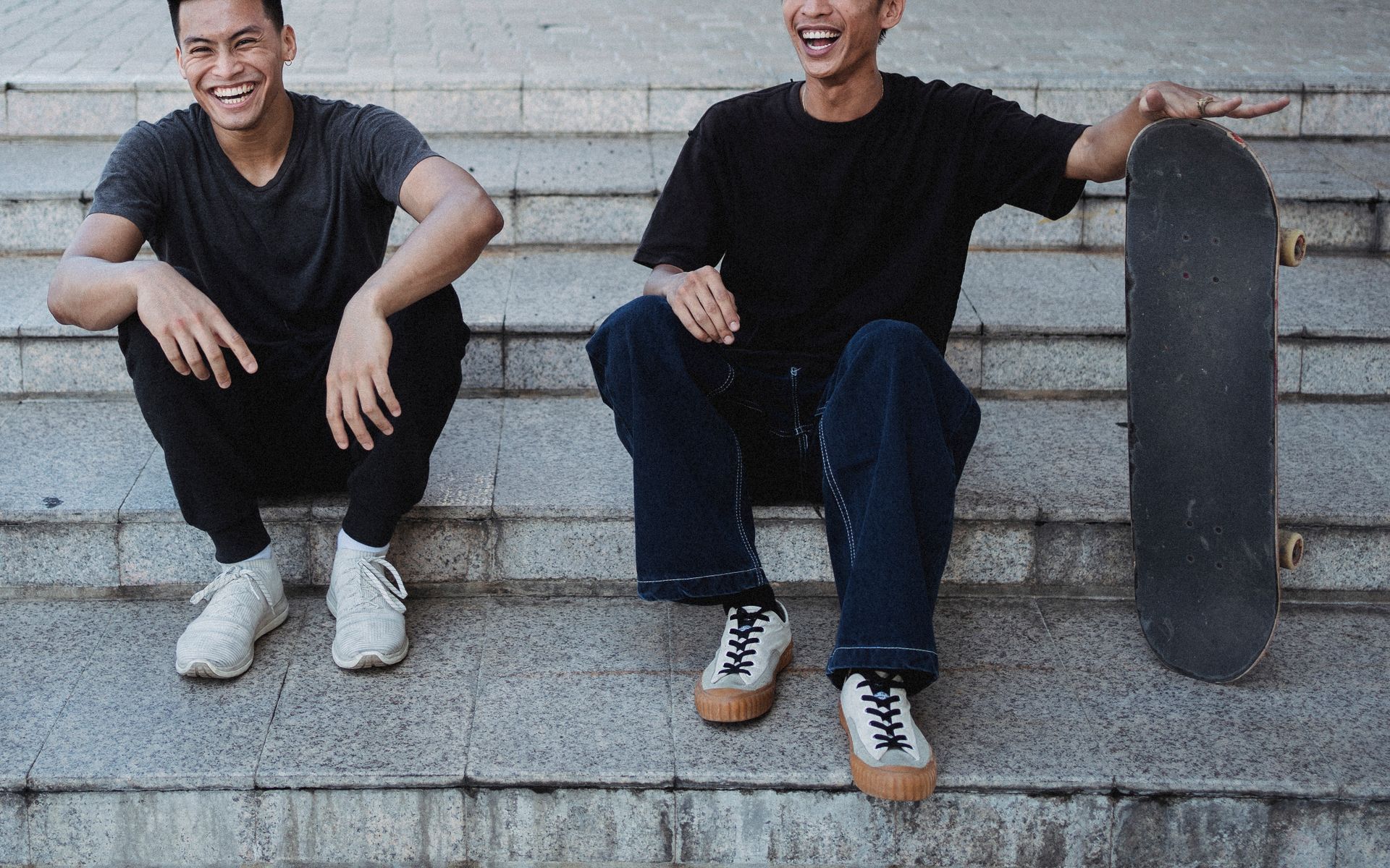 Two cheerful young men with a skateboard sitting on steps outdoors.