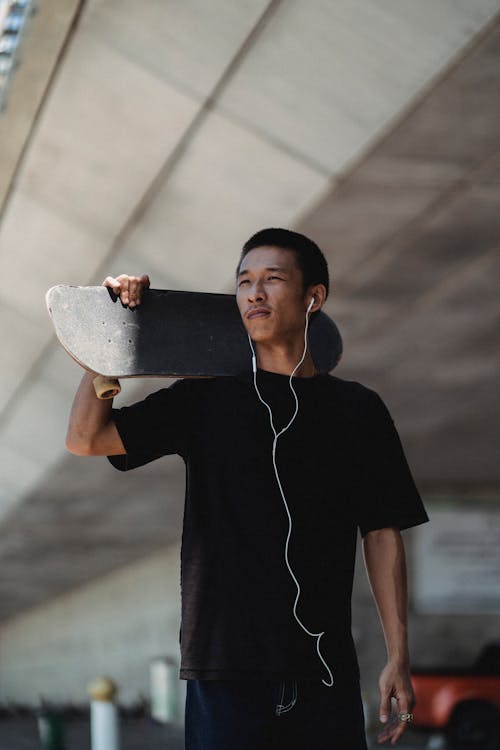 Positive Asian skater in earphones carrying skateboard on shoulder