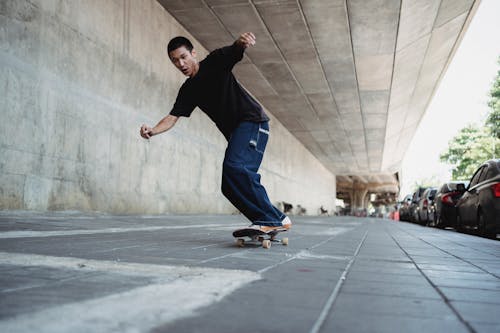 Full body positive Asian male in dark casual outfit riding skateboard along paved pedestrian walkway on sunny weather