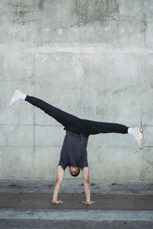 Full body anonymous flexible male in activewear performing handstand and doing split in air against shabby concrete wall