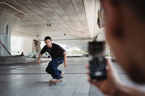 Free Unrecognizable person taking picture on smartphone of Asian male friend riding skateboard under transport bridge in city center Stock Photo