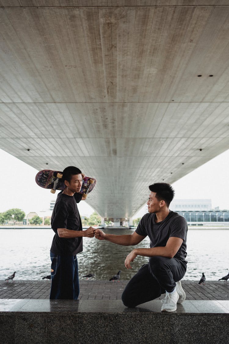 Smiling Asian Men Giving Fist Bump On River Embankment Under Bridge