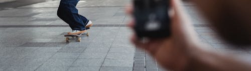 Faceless person taking picture on cellphone of unrecognizable person in white sneakers riding skateboard along pavement