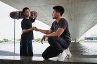 两个年轻人在城市河边的桥下玩滑板，握手致意，洋溢着友谊的氛围