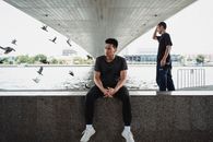 一位身着黑衣、脚穿白色运动鞋的亚洲人神情严肃地坐在石堤上，他的朋友站在桥下，周围有鸽子飞过