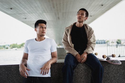两个年轻人拿着滑板，坐在桥下，享受户外的轻松时刻