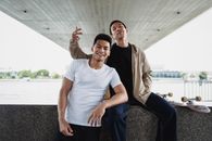Positive young male friend leaning on knee of friend on river embankment in city