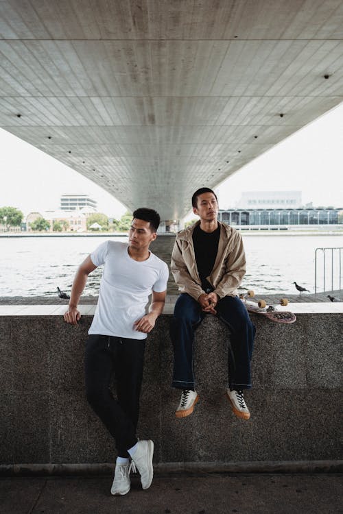 Confident Asian male leaning on granite fence next to sitting with skateboard young brother under wide river bridge in megapolis