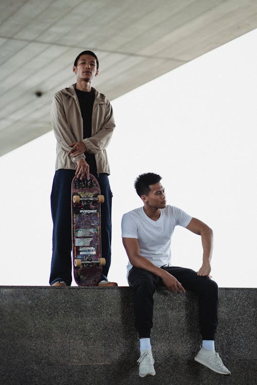Full body of tranquil Asian men with skateboard standing and looking at camera sitting on steps in daytime