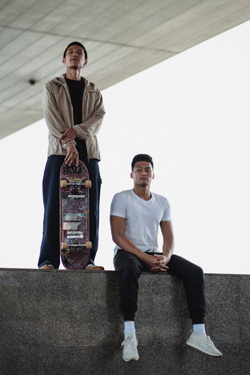 Calm man with colorful skateboard standing next to serious man in sportswear sitting on steps