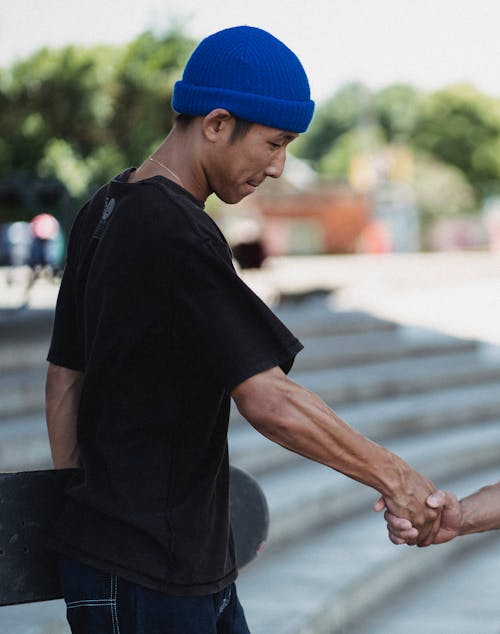 Side view of glad skater in blue cap and black shirt shake hands with man in skate park in daytime