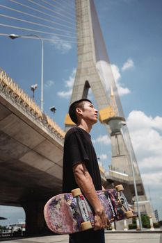 晴朗的天空下，一个背着滑板的年轻人站在吊桥旁，若有所思