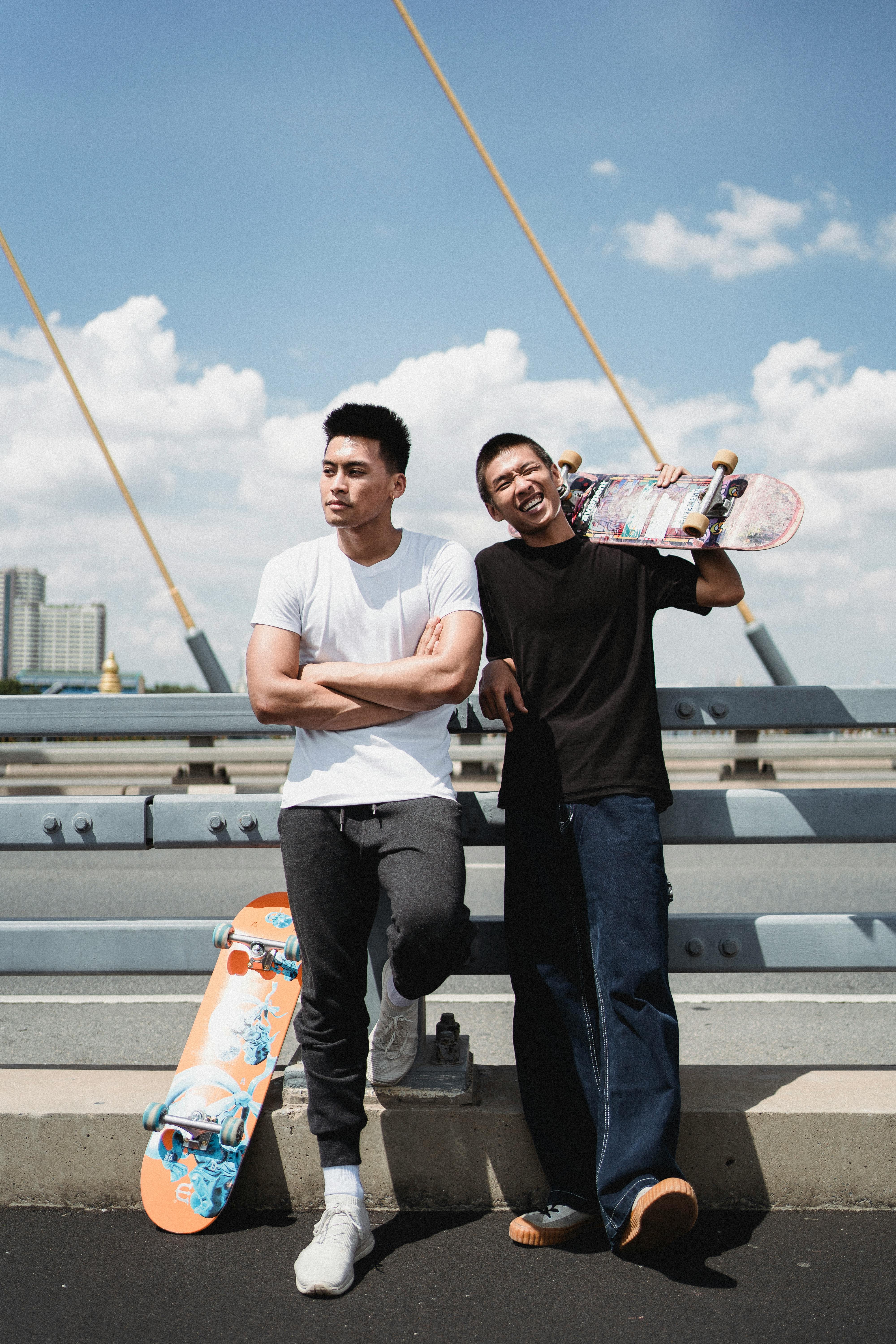 年轻的民族运动员站在镇上的桥边，身旁是带着滑板的梦幻男性伴侣
