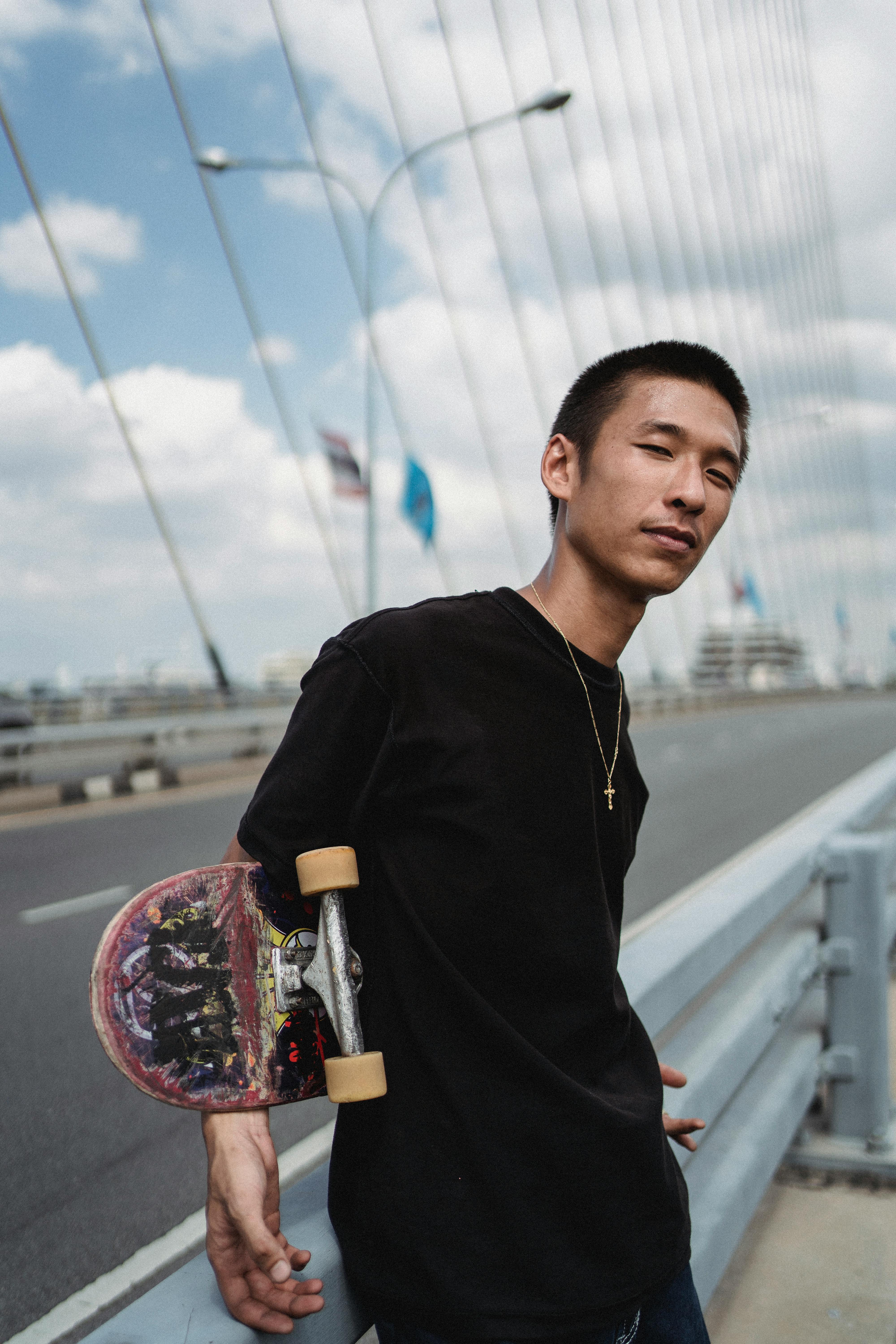 年轻的民族运动员拿着滑板，在多云的天空下，于城市道路附近注视着摄像机