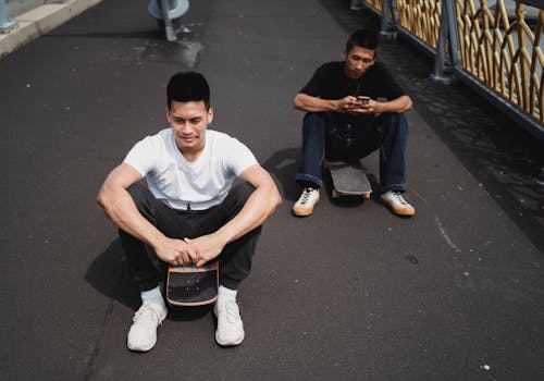 Young content ethnic sportsman resting on skateboard near male partner surfing internet on cellphone on pavement